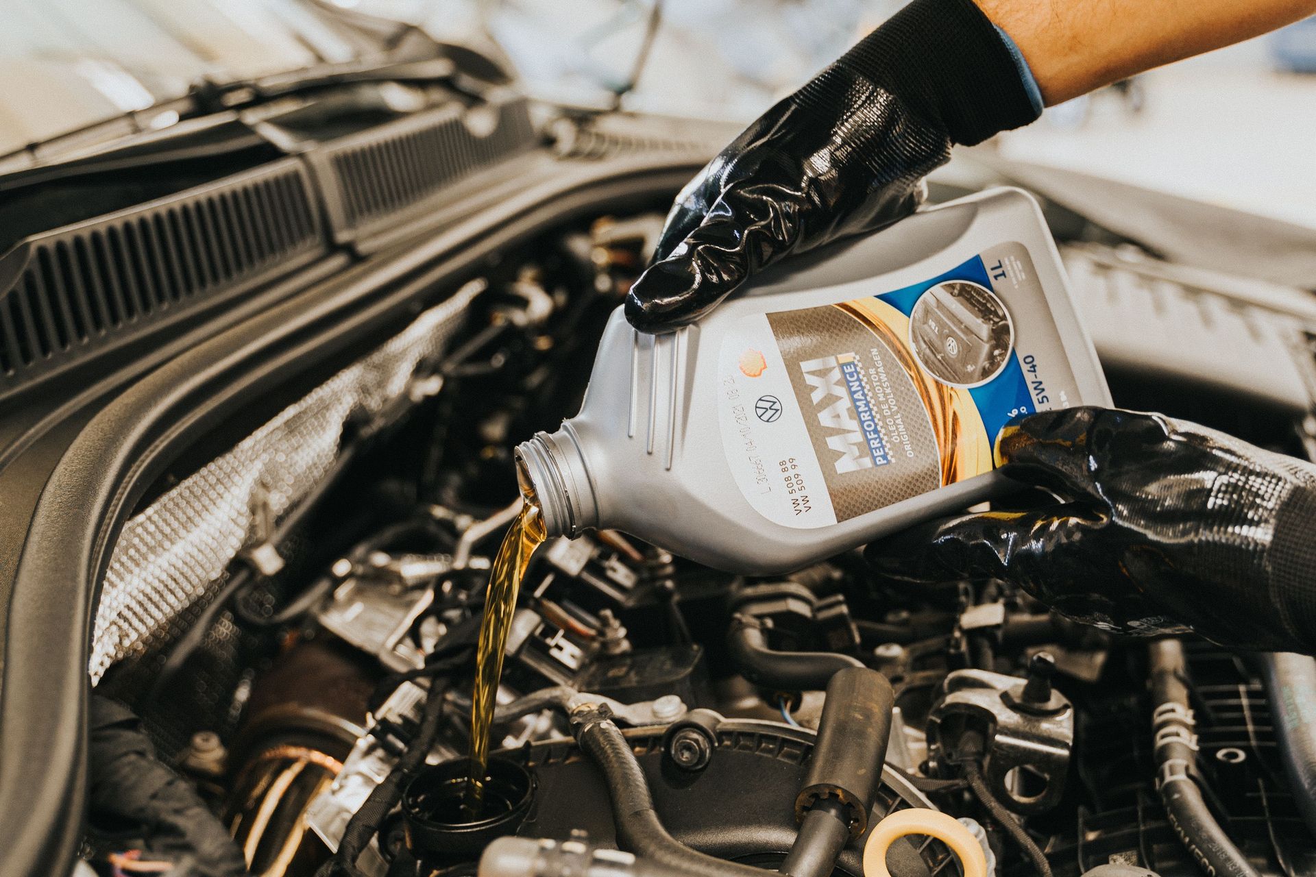 A mechanic wearing gloves pouring oil into a car engine in a garage setting.